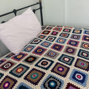 May include: A colourful crocheted throw blanket with a granny square pattern in shades of blue, purple, pink, red, and green. The blanket is made of soft yarn and has a white background.