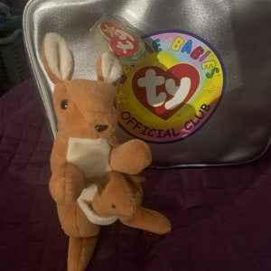 May include: A brown kangaroo Beanie Baby with a joey in its pouch. The kangaroo is sitting on a purple surface. The Beanie Baby is in a silver Ty Beanie Babies bag with a red Ty tag.