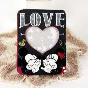 May include: Black picture frame with a heart-shaped cutout featuring silhouettes of Mickey and Minnie Mouse. The word "LOVE" is written in white. The frame has red and yellow accents and is placed on a cream-colored crocheted blanket.