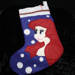 May include: A blue Christmas stocking featuring a cartoon image of a mermaid with red hair and blue eyes. The stocking has white polka dots and a white cuff with red trim. A candy cane is placed on the top.