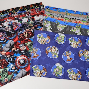 May include: A collection of rectangular fabric items with various colorful patterns. Designs include comic book superheroes, and characters from the movie Toy Story. The items are likely placemats or similar home decor items.