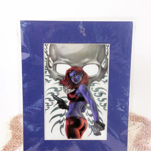 May include: Comic art print featuring a female character with blue skin, red hair, and a red top, holding two guns. The artwork is framed in a blue mat, with a skull in the background.