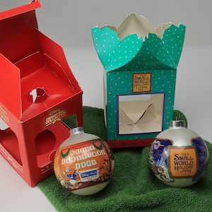 May include: Two glass Christmas ornaments with Disney character designs, next to their original boxes. One ornament has a design with Donald Duck and the text "Disney's Christmas Collection". The other ornament has a design with the text "It's a Small World Holiday 1994".