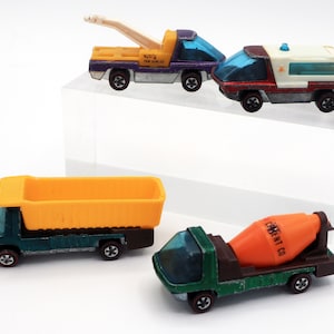 May include: Four vintage toy trucks in various colors. The collection includes a yellow tow truck, a white ambulance, a green dump truck with an orange bed, and a green cement mixer truck. Each truck has a clear, bubble-like top and red-lined wheels.