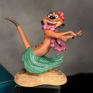 May include: A figurine of Timon from The Lion King, posed in a hula skirt and lei. The figure is tan and brown with a green skirt and pink lei. The base is a sandy color.