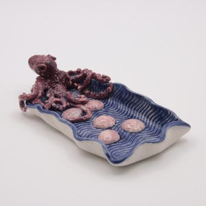 May include: A ceramic decorative tray with a marine theme. The tray is shaped like a wave, with a blue and white color scheme. It features a purple octopus and several pink seashell-shaped objects.