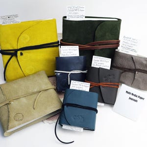May include: A collection of handmade leather journals in various colors, including yellow, green, blue, and gray. Each journal is bound with a leather cord closure. Some have attached tags with handwritten text. The journals are labeled "Multi-Media Paper Journals."
