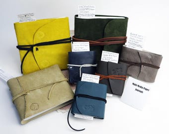 May include: A collection of handmade leather journals in various colors, including yellow, green, blue, and gray. Each journal is bound with a leather cord closure. Some have attached tags with handwritten text. The journals are labeled "Multi-Media Paper Journals."