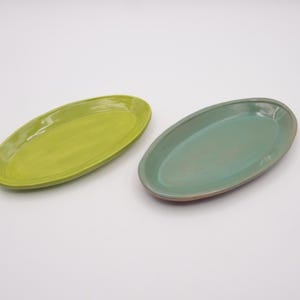 May include: Two oval-shaped ceramic serving platters. One is a bright lime green, the other is a muted teal. Both have a smooth, glossy finish and are set against a white background. These platters are ideal for serving appetizers or small dishes.