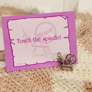 May include: A pink card with the text "Touch the spindle!" and a silver spinning wheel charm. The card is set on a crocheted blanket with pink and cream colors. The image has a fairytale theme.