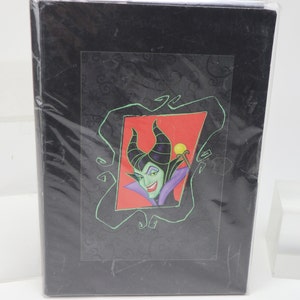 May include: A black book or journal wrapped in clear plastic. The cover features a framed image of a green-skinned woman with black horns, a purple collar, and a red background.