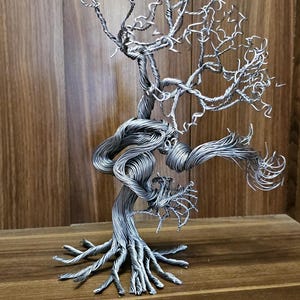 May include: A detailed wire sculpture of a tree. The silver wire is carefully shaped to create the trunk, branches, and roots. The sculpture is displayed on a wooden surface, with the tree measuring approximately 25 cm in height.