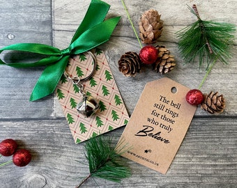 May include: A silver jingle bell keychain with a green ribbon bow. The keychain is on a card with a Christmas tree pattern and the text "The bell still rings for those who truly Believe" on a brown tag.