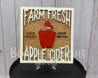 May include: A wooden sign with white lettering that reads "Farm Fresh" and "Apple Cider". The sign also features a red cutout of a jug with a twine bow. The text "Hand Picked" and "Home Grown" are also on the sign.