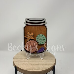 May include: A wooden jar-shaped sign with a brown background and a silver outline. Inside the jar are colorful wooden cutouts of a starfish, a seashell, and a snail shell. The sign is displayed on a small wooden stand.