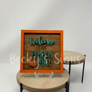 Puede incluir: Un letrero de madera con un marco naranja y un fondo de madera. El letrero dice "Bienvenido a nuestro huerto de calabazas" en letras color turquesa con un gráfico de calabaza turquesa.