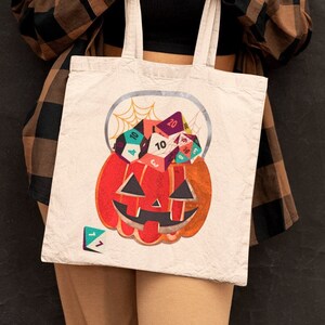 May include: A white tote bag featuring a jack-o'-lantern design filled with colorful dice. The pumpkin is orange with a carved face, and a spiderweb is above it. The dice are various colors and shapes, perfect for Halloween or tabletop gaming.