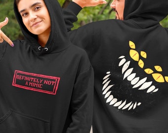 Puede incluir: Sudadera con capucha negra con bolsillo delantero. La parte delantera de la sudadera tiene el texto "DEFINITELY NOT A MIMIC" en un rectángulo rojo. La parte trasera de la sudadera presenta un gráfico de monstruo blanco y amarillo.
