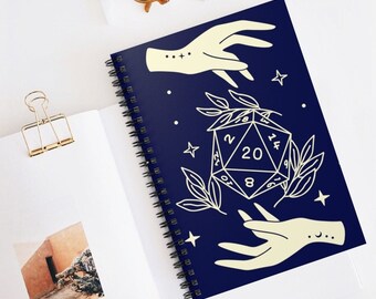 May include: A dark blue spiral notebook with a white illustration of a 20-sided die, two hands reaching towards it, and stars and leaves. The die has the numbers 2, 4, 6, 8, 10, 12, 14, 16, 18, 20, 1, 3, 5, 7, 9, 11, 13, 15, 17, and 19.