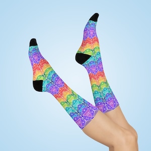 May include: A pair of colorful socks with a rainbow pattern. The socks feature a repeating design of twenty-sided dice in various colors, with black heels and toes. The socks are displayed against a light blue background.