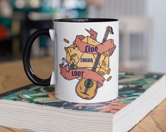 Pode incluir: Caneca de cerâmica branca com alça preta. A caneca tem um gráfico de um dado com uma fita que diz "Live Laugh Loot" e um alaúde. O gráfico está em tons de amarelo, laranja e marrom.