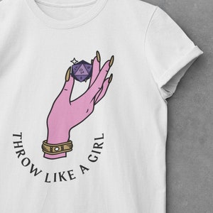 Puede incluir: Camiseta blanca con una mano rosa sosteniendo un dado morado de 20 caras. El texto "Throw Like A Girl" está impreso en un círculo alrededor de la mano.