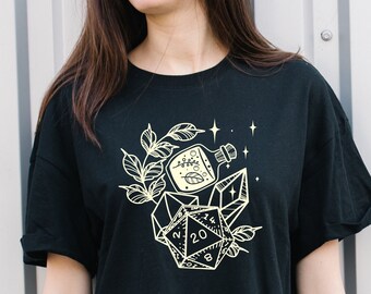 Puede incluir: Camiseta negra con un diseño gráfico blanco de un dado de 20 caras, una botella y una rama con hojas. El diseño está rodeado de pequeñas estrellas.