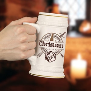 Puede incluir: Jarra de cerveza de cerámica blanca con borde dorado y diseño marrón con un dado de 20 caras, espadas cruzadas y el texto "Christian".