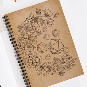 May include: Spiral-bound notebook with a kraft paper cover. The cover features a black line drawing of flowers and geometric shapes. The notebook is open, revealing white pages. Sunglasses are visible in the background.