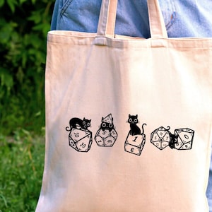 Può includere: Una borsa tote in tela bianca con un'illustrazione in bianco e nero di gatti seduti su dadi. I dadi mostrano i numeri 1, 2, 6 e 20.
