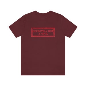 May include: Burgundy t-shirt with a red graphic that says "DEFINITELY NOT A MIMIC" in a vintage-style stamp.