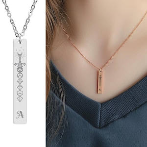 Peut inclure: Un collier argenté avec un pendentif rectangulaire. Le pendentif présente un motif gravé d'une épée, de dés et d'autres symboles, avec la lettre "A" en bas. Le collier est présenté sur fond blanc.