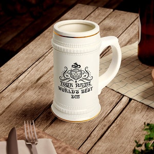 Könnte beinhalten: Ein weißer Keramik-Bierkrug mit goldfarbenem Rand und einem schwarz-weißen Design mit einem Drachen, einem zwanzigseitigen Würfel und dem Text "Your Name World's Best DM".