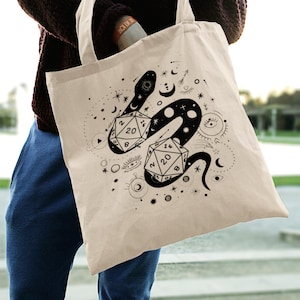 May include: A beige tote bag with a black and white design featuring a snake wrapped around a twenty-sided die. The design also includes stars, moons, and other celestial symbols.