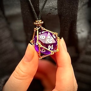 May include: A purple 20-sided die with gold wire cage, hanging from a black cord with a gold clasp. The die is hanging from a person's hand.