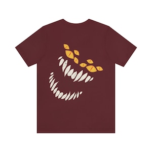May include: A maroon t-shirt with a white and yellow graphic of a monster's face with sharp teeth and multiple eyes.