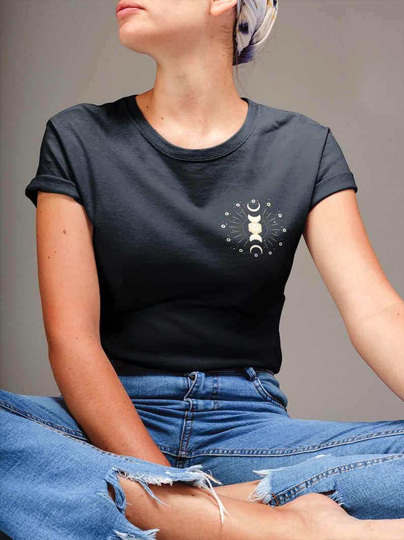 May include: A dark gray t-shirt with a white moon phases design on the left chest. The design features a crescent moon, a full moon, and a half moon with a sun in the center. The shirt is worn by a person sitting with their legs crossed.
