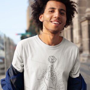 May include: A white t-shirt featuring a black and white illustration of a hand holding a twenty-sided die. The die is floating above the hand. The text "YEET" is printed below the illustration.