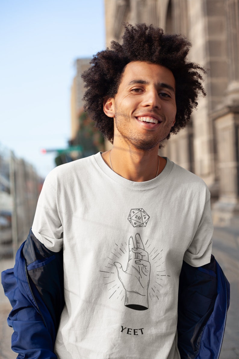 May include: A white t-shirt featuring a black and white illustration of a hand holding a twenty-sided die. The die is floating above the hand. The text "YEET" is printed below the illustration.