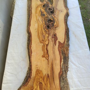Op de afbeelding: Een houten plank met natuurlijke rand en een schorsrand. De plank heeft een lichtbruine kleur met donkerbruine en roodbruine nerfpatronen. Het hout heeft verschillende knopen en onvolkomenheden.