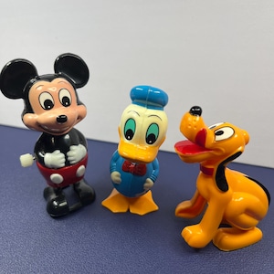 May include: Three vintage Disney character figurines: Mickey Mouse, Donald Duck, and Pluto. Mickey is in black, red, and white. Donald Duck is blue, yellow, and white. Pluto is yellow and black. All are standing on a blue surface.