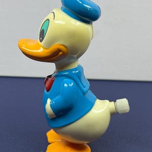 May include: A vintage Donald Duck wind-up toy. The toy features a yellow body, orange beak and feet, and is dressed in a blue sailor shirt and hat with a red bow tie. The toy is a classic collectible.