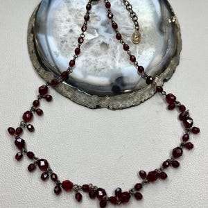 May include: A delicate red glass bead necklace with a dangling design. The necklace is made with small, faceted beads and has a simple clasp.