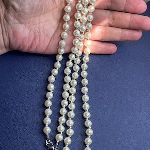 Pode incluir: Um colar de pérolas de várias voltas com fecho prateado. O colar apresenta pérolas redondas, esbranquiçadas, separadas por pequenas contas prateadas. O fecho tem um pequeno pingente decorativo. O colar é exibido sobre um fundo azul escuro.