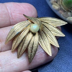 Puede incluir: Broche dorado con diseño floral, con pétalos texturizados que irradian de una perla central. El broche tiene una superficie detallada y en relieve, y lo sostiene una mano sobre un fondo azul. Es una joya vintage.