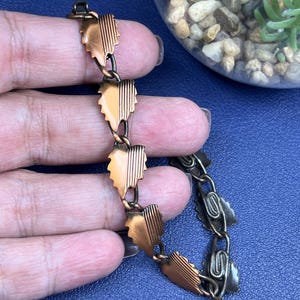 May include: A vintage copper bracelet featuring leaf-shaped links. Each leaf has a textured, ridged design and is connected by small, dark metal links. The bracelet is held against a blue background, showcasing its intricate details and warm tones.