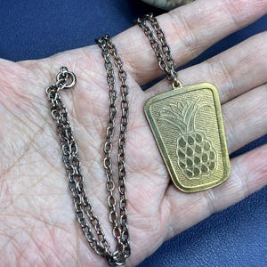 May include: A vintage gold-tone necklace featuring a rectangular pendant with a pineapple design. The pendant hangs from a delicate chain. The necklace is displayed on a hand against a dark blue background.