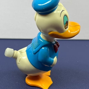 May include: A vintage Donald Duck wind-up toy. The toy features a blue sailor hat, jacket, and bow tie, with a white body, orange beak and feet. The toy is in a walking pose, with a wind-up key on its back.