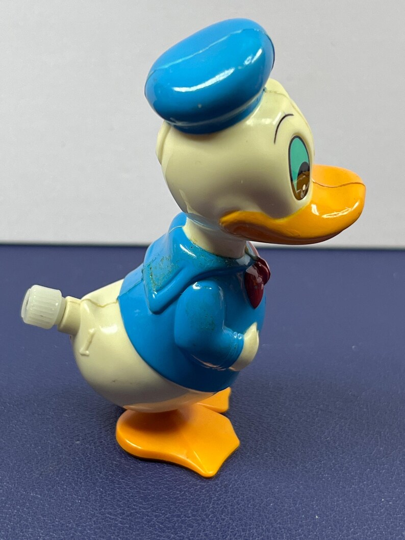 May include: A vintage Donald Duck wind-up toy. The toy features a blue sailor hat, jacket, and bow tie, with a white body, orange beak and feet. The toy is in a walking pose, with a wind-up key on its back.