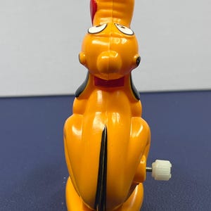 May include: A vintage Pluto wind-up toy. The toy is primarily orange with black ears and a black stripe down its back. It has a red bow tie and a white crank on the side. The toy is a collectible item.